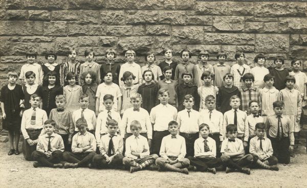 Students pose outdoors in front of a stone wall at Saint Raphael's School. Mary Julia O'Dea is standing in the 3rd row, first on the right. She is possibly in the 5th grade.