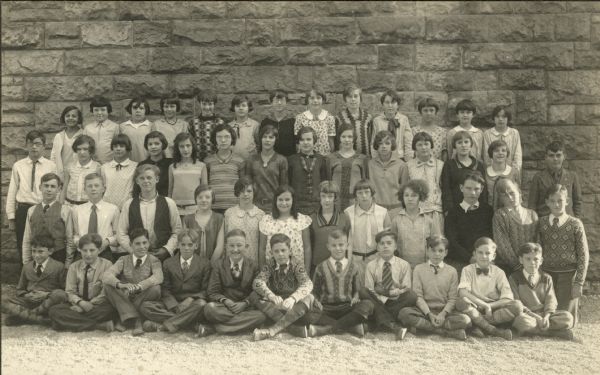Students pose outdoors in front of a stone buildings at Saint Raphael's School. Mary Julia O'Dea (born 1917) is standing in the third row, third from the left. She is in the 7th grade.