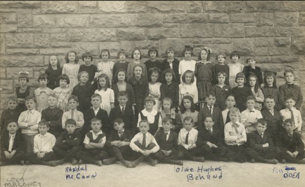 Students pose outdoors in front of a stone buildings at Saint Raphael's School. Bill O'Dea is sitting cross-legged in the 1st row, first on the right. Randal McCann may be the boy seated in the 1st row, either 3rd or 4th from the left. Olive Hughes Behrnd is kneeling in the 2nd row, 8th from the left. Lela Maloney may be standing in the 3rd row, 4th from the left. This class is the 4th or 5th grade.
