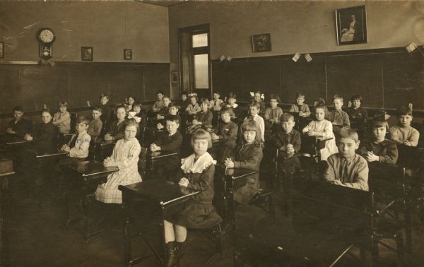 Class portrait at Pettit School, also known as Saint Raphael's School. Thomas O'Dea is sitting in the 1st row of desks, 3rd from the left. All of the students are sitting up very straight with their hands folded on their desks. Blackboards decorated with small American flags cover the walls in the background, and a clock can be seen above on the left. The door is in the back corner.