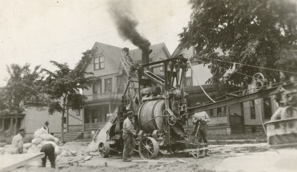 Cement mixer and construction crew at a work site. Smoke pours from the exhaust pipe on top. Bags of material and other equipment can be seen on the left and right. Houses and trees are in the background.