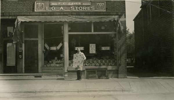View from street of a man standing on the sidewalk in front of a grocery store at 300 West Lakeside Street. The overhead sign reads, "F.C. Caves, Meats and Groceries, Member, I.G.A. Stores" with two IGA signs on the right and left. He is holding a sign covered with large polka dots. A smaller, similar sign is being held at an angle over the larger sign. The sign is probably part of a promotion for the South Side Picnic being held on Sunday, June 14th, 1931. Groceries and their prices are displayed in the windows. A Hills Brothers Coffee sign is next to the door. The store has a crank out awning above the display windows.