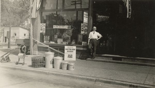 Henry Esser stands on the step in front of Koltes & Esser Hardware in the 300 block of West Lakeside Street. Fencing, garbage cans, a hose reel and lawn mower are on display at the curb. The rear of the company truck can be seen parked on the left. The window is full of merchandise and signs. Bamboo fishing poles are stacked diagonally across the front. A sign for the South Side Picnic to be held on Sunday, June 14th, 1931, is propped against the store foundation. It reads "Dance, South Side Picnic, Sun. June 14, Olin Park, Board Compliments, Capital City Lumber Company." Many of the South Madison businesses displayed similar signs.