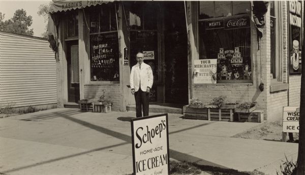 View from street of Saul Kasdin posing in front of Kasdin's Fancy Groceries & Meats, a store at 402 West Lakeside Street. Merchandise and prices are displayed in the window, "Pot Roast 17¢ per #" and "Tomatoes 2# for 19¢" and "Red Top Malt Syrup." A Schoep's Home-ade Ice Cream sign stands on the terrace. Flats of plants (probably vegetables) are stacked on crates against the foundation. In the window on the right, is a sign, with food items attached, for the South Side Picnic to be held on Sunday, June 14th, 1931. It reads, "Your Merchants Share With U, Trade with them, Board Compliments, Capital City Lumber Company."