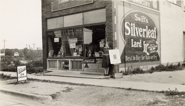 View from street of a woman standing at the corner of the Lakeside Grocery, located at 109 East Lakeside Street. On her left, a sign is propped on a crate for the South Side Picnic to be held on Sunday, June 14th, 1931. It reads, "Plan To Attend, June 14th, Hear the Band, South Side Picnic." The store has merchandise and signs in the windows. Near the street is a "Kennedy's Velvet Ice Cream" sign. On the side of the building is a large painted advertisement for "Swift's Silverleaf Lard, Best to Buy for Bake or Fry."