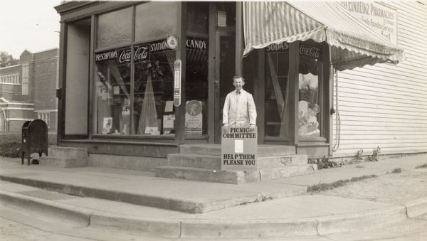 Charlie Collins of the Kleinheinz Pharmacy stands on the stoop holding a sign for the South Side Picnic to be held on Sunday, June 14th, 1931. It reads, "Picnic Committee, (followed by a list too small to read), Help Them Please You, Board Compliments, Capital City Lumber Company." The location of the pharmacy was 335 West Lakeside. Merchandise and signs are on display in the windows. The business included a soda fountain and a pay phone. A post box and Franklin Elementary School can be seen on the left. There was another Kleinheinz Pharmacy on Park Street.