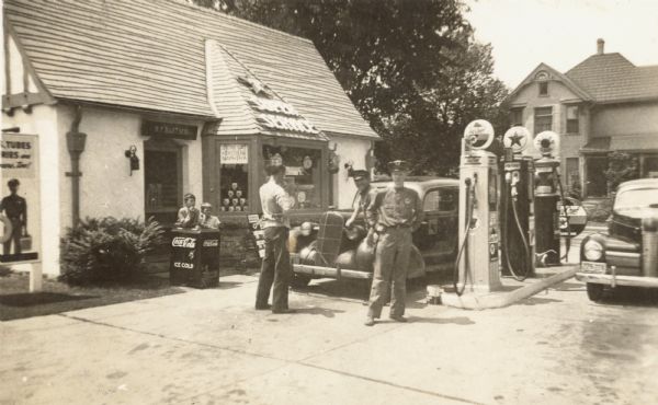 A Texaco Super Service filling station owned by H.F. Bartsch on the corner of Park and Midland Streets. A home is on the opposite side of Midland Street. Two boys are leaning on the Coca Cola cooler. Cars are parked on each side of the gas pumps. Two attendants pose in uniform while another man stands with his back to the camera. The glass globes on top of the pumps read "Sky Chief," "Texaco" and "Indian Gas." A sign is the window advertises "Stove Gas, Kerosene, Naphtha."
