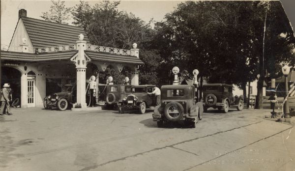 Leyda's Service Station on the corner of Park Street and Olin Avenue. This service station was a member of the Wadham's Oil and Grease Company chain of Milwaukee, Wisconsin. They built distinctive pagoda-like buildings influenced by Japanese culture. Five automobiles can be seen driving in, out and being serviced by uniformed attendants. Two people are at the far left. Olin Avenue, lined with trees and homes is in the background. Handwritten caption below, "Leyda's Station, 40, 24 Hour Servis [sic]."