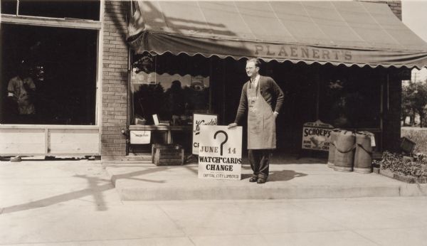 Walter Plaenert, owner of Plaenert's Grocery, poses in front of his store at 1128 South Park Street. He is holding a sign for the South Side Picnic. The sign reads "June 14, Watch Cards Change, Board Compliments, Capital City Lumber Company." It also has a large question mark in the center. The store has merchandise in the windows and signs for "Kennedy's Creamery," "It's a Food, Schoep's, Home-ade Ice Cream," "Lake Shore Seeds," "Central Wisconsin Co-operative." On the right, vegetables in flats are for sale. On the left is a window with a workman in an apron peering out. His apron is advertising a lumber company. There is a building permit below Plaenert's window.