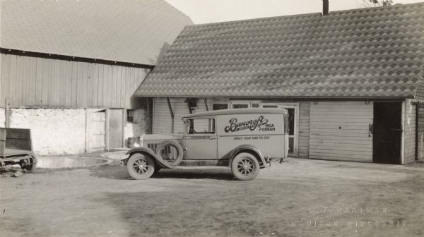 View across yard of a Bancroft Brothers delivery truck parked near the corner of a barn and shed. The back of a parked wagon is on the left, near two large round concrete pits in the ground. Text on the truck reads, "Bancroft Brothers, Milk and Cream," "Direct From Farm To You" and "Oakwood 88-J-12."