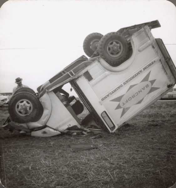 An upside down Bancroft delivery truck. A man is standing just behind the front tires. On the right, a "Dane County Police Traffic" car can be seen behind the truck box. Text on the truck reads, "Bancroft (inside an oval with an eight point star), From America's Dairyland." The reason the truck is upside down is unknown.