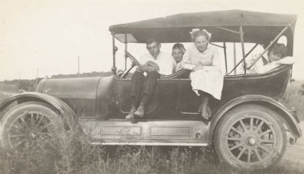Three O'Dea sisters and an unidentified man pose while looking out the left side of an automobile. The automobile has a fabric roof but no windows. A dirt road is just behind them.