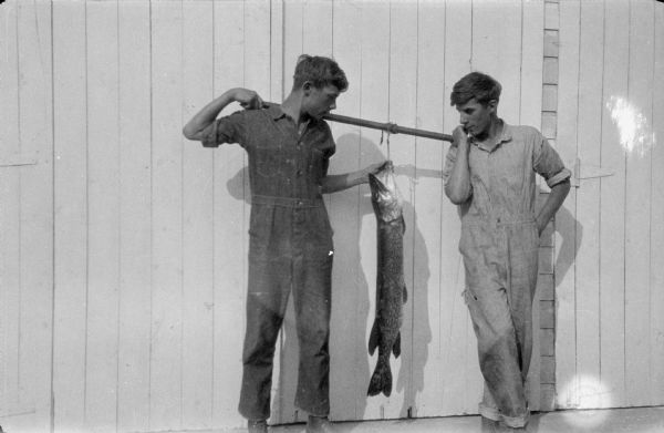 Tom and Bill O'Dea pose with a large Northern Pike in front of their parent's garage door on Park Street. They are wearing one piece coveralls and boots. The fish is suspended between them hanging on a pole.