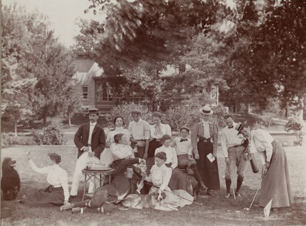 An outdoor family portrait with a golf theme. Several members are sitting on a bench and the lawn, with a large house in the background. There are two dogs, one in the center front, one on the left near a woman holding a ball. Refreshments sit on a table, and a reclining man is pouring liquid into a glass for a woman seated next to him.