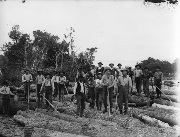 Driving crew posing on the logs. They are holding peaveys and cant hooks, tools used to move logs. The man on the left is holding a pail and the man in the center is holding a pail in one hand and a bowl in the other. Trees are in the background.