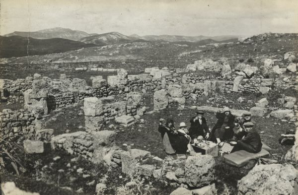Slightly elevated view of a group of men, women and children having a meal outdoors on the ground. They are gathered around a box being used as a table. Cushions are placed on the rocks and grass to serve as seats. They are saluting the photographer with their coffee cups. All around them are ruins of an archaeological site. Mountains and hills are in the distance. Kremassa was an old Roman city near Souk Ahras, Algeria.