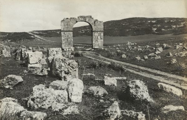 The remains of an arch surrounded by tumbled stones and ruins of an archaeological site. A dirt track in the foreground goes through the arch and rises to hills in the distance. Kremassa was an old Roman city near Souk Ahras, Algeria.
