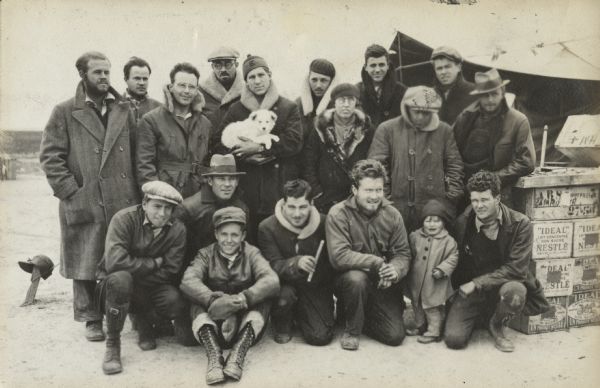 Outdoor group portrait of the Logan African Expedition, Berriche, Algeria. Six men and a toddler are crouching or kneeling in the foreground. Behind them men, and one woman, are standing in two rows. The man standing in the center is holding a small white dog in his arms. On the right is a tent and a stack of boxes of supplies. A tent stake with a hat perched on it is on the left. Right to left, back row: Voight, Krieger, Small, Greenlee, Sharp. Second row, Williams, (Alonzo) Pond, Dorothy Long Pond, Roberts, Moen, Brown: Bottom row, Riedel, Chomingwen (daughter of the Ponds), Gillin, Tax, Wilford, Dobson (front) Nash. They are wearing work clothes, overcoats, hats and boots.