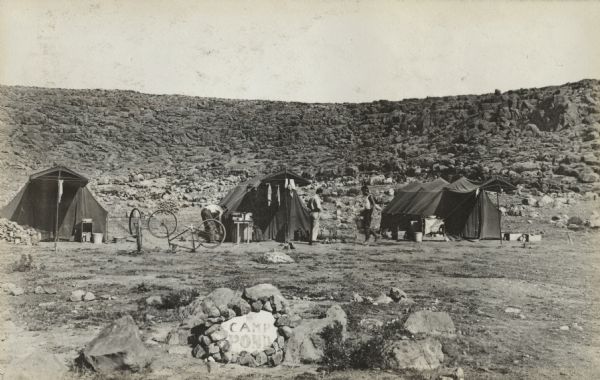 Camp Pond in North Africa. A rock in the fire pit in the foreground has "Camp Pond" painted on it. Three tents, three people, two upside down bicycles and various camp objects are just beyond the fire pit. Rock covered hills are in the background.