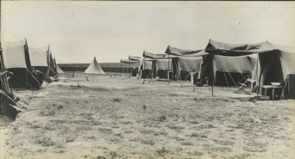 The back row at Camp Logan. Ten large tents, some with awnings, are lined up facing each other, five on the right and five on the left. The tent in the center background is of the center pole style. Table, chairs and other camp equipment are under the awnings.