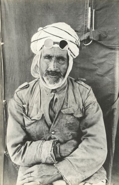 A man named Chabane is posing while sitting in front of a tent. He is wearing a work jacket and traditional headdress, with goggles pushed up on his forehead. His arms are crossed on his lap. He has a beard and moustache.