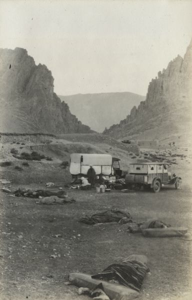 The camp at El Kantara, Algeria. In the foreground, members of the expedition are sleeping on pallets, under blankets. Just beyond the sleepers are two trucks and two men with camping equipment, pails and tubs. Between the trucks two cots also appear to have sleepers in them. In the middle ground are stone walls and structures. The gorge seen in the background has been described by locals as "Mouth of the Desert." Mountains are in the far distance.