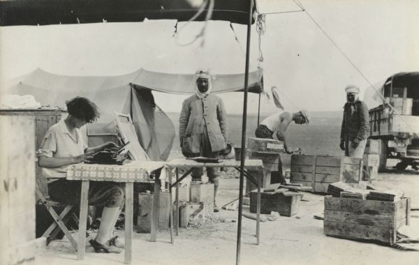 A woman is working on a Remington typewriter, set on a table with a patterned tablecloth, on the last day in camp. One man is standing in the center, and two more men are packing a crate on the right, next to a truck. All the men are wearing goggles. Two tents are still pitched and more boxes are being filled. More camp equipment and fuel or water cans are piled near the tents.