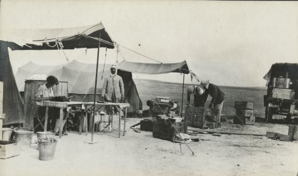 A woman is recording the contents of each box on a Remington typewriter, set on a table with a patterned tablecloth, on the last day in camp. Three men are standing in the center, two of them packing a crate. Two tents are still pitched and more boxes are being filled. More camp equipment and fuel or water cans are piled near the tents.