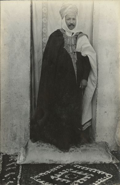 Caid Kahlifa Lamin poses in front of an opening, perhaps a door, in a cement wall, covered with a cloth hanging. He is dressed in official attire and is standing on a cushion placed on a carpet.