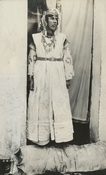 Dorothy Pond posing dressed in indigenous apparel, including jewelry, as a Mauresque. She appears to be standing in a doorway partially covered with a cloth hanging, with a cushion at her feet.