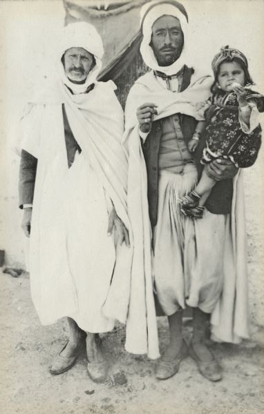 Two indigenous men are posing for the camera in Ain Beida, Algeria. One man is holding a young boy in his left arm.
