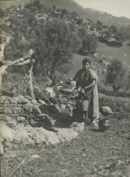 A Kabyle girl holding a pail at the well, with another pail sitting on the ground at her feet. The well has a stone wall around it with a rope and pulley suspended from a wooden frame. A tree-covered slope and mountains are in the background.