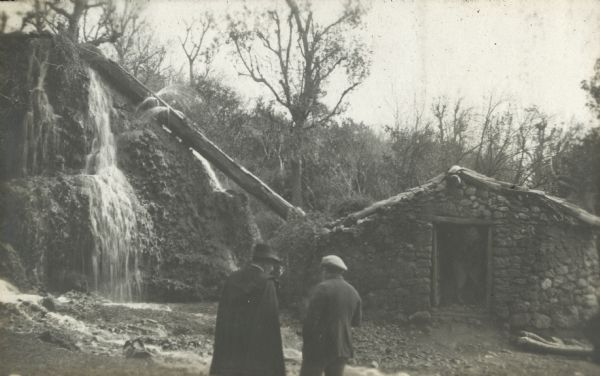 Two men are standing in front of a stone mill house and wooden flume. Another man is just visible crouching inside the doorway of the mill house. The flumes that carry water to the mill house are made of hollow tree trunks. This mill grinds oats and wheat. Trees are in the background.