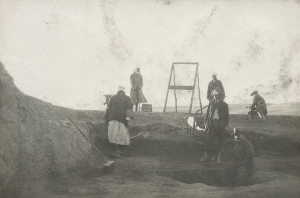 Workmen excavate at an archaeological site at Mechta. M. Bebruge is the man in the foreground. A man sitting on the ground in the background on the right is leaning towards a dog.