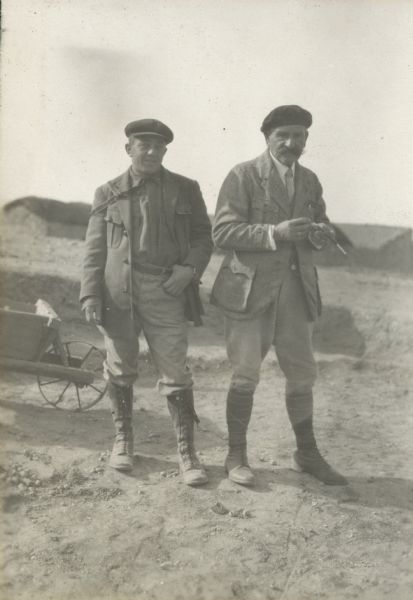 Alonzo Pond and colleague, M. Debruge, posing for a photograph at Mechta. M. Debruge has a cigarette in a holder in his hand, and both men are wearing coats, hats, jodhpurs and tall lace-up boots. The front of a wheelbarrow is on the left just behind the two men. Buildings are in the background.