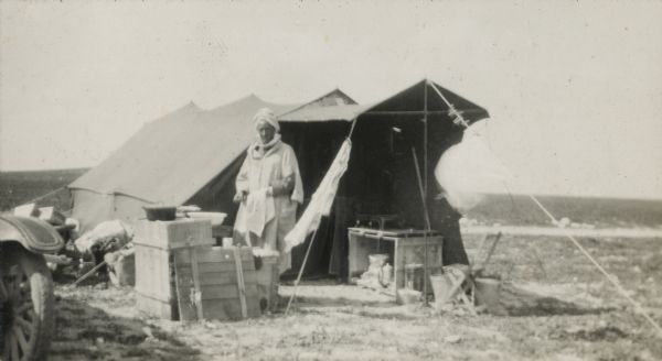 Ferrah stands in front of a tent in the Pond camp. Boxes are being used as tables, chairs and other camp supplies aresitting on the ground near the entrance, and along the side. Part of a wheel and fender of what may be a truck is on the left. The desert landscape stretches towards the horizon.