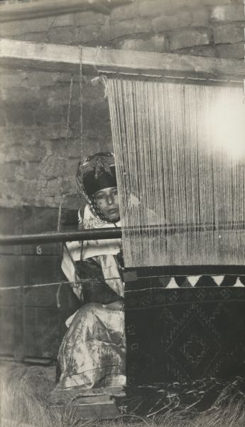 A woman is sitting at a loom, weaving a rug.