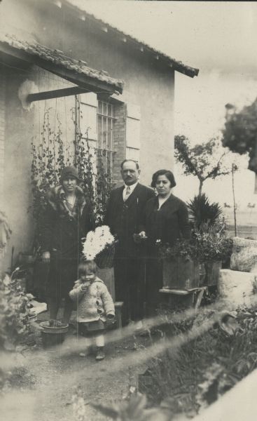 Dorothy Pond and her daughter, Chomingwen Pond, pose with Monsieur and Madame Bernard in front of their house in Canrobert, Algeria. Flowers are planted in containers and vines are trained on strings against the wall.