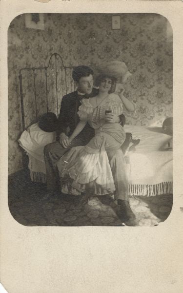 A gag photo postcard of a man, seated on the edge of a bed, with his arm around a life-sized paperboard cut out of a beautiful, seated woman. He is holding her hand with his right hand, and he holds a glass in his left hand with his arm around her waist. He is wearing a suit and eyeglasses, and his hat is on the pillow to his right. She is wearing a dress and feathered hat, her legs are crossed, and she is holding up a glass near her face. The bed has a metal headboard and a light-colored bedspread with fringe. The walls are covered with wallpaper, and two framed prints are hanging on the wall. A carpet covers the floor.