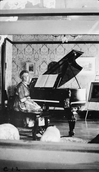 Interior view of home, perhaps taken through an open window. The sash and sill are in the foreground. A woman is sitting on the piano bench in front of a grand piano. On the far right a painting sits on an easel.