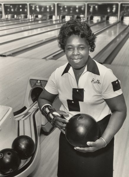 A woman smiles for the camera as she poses with her bowling ball. The caption reads, "Bertha Eichelberger bowls in five leagues every week." She is wearing a bowling shirt with "Berth" embroidered on it and a brace on her right wrist. The bowling lanes and ball return machine are in the background.