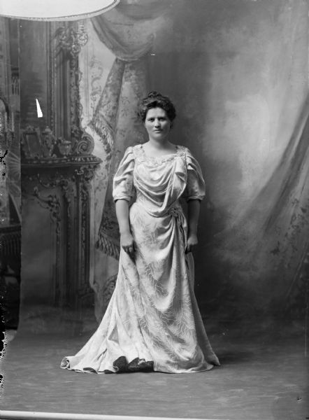 Formal, full-length portrait in front of a painted backdrop of Belle Case La Follette, the wife of Robert M. La Follette, Sr. Although the negative jacket identified the portrait as 1903-1904, the La Follette Family album, which was assembled by Isabel La Follette, identifies it as 1905-1906, which seems more likely. It is also likely the dress is a new one purchased to wear in Washington, D.C.