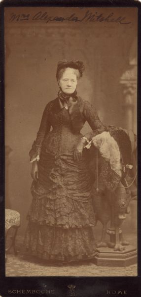 Cabinet card of a full-length studio portrait of Mrs. Alexander Mitchell standing in front of a painted backdrop. She is wearing a floor-length dark-colored dress, leather gloves and a bonnet tied under her chin with a wide ribbon. She is resting her arm on a claw footed stand on which is draped a furred coat or cape.<p>Martha (Reed) Mitchell (1818-1902) was born in Westford, Massachusetts, the daughter of Seth Reed. The family moved to Milwaukee in 1836 or 1837. She married Alexander Mitchell (1817-1887, bn. Scotland) in 1841. Alexander moved to Milwaukee in 1839, and eventually became a millionaire from his banking and railroad concerns, specifically the Milwaukee & St. Paul Railway. He also served two terms as a US Representative from 1870 to 1874. Mrs. Mitchell was one of the founders of the Milwaukee Museum of Fine Arts in 1882 and the Woman's Club of Wisconsin in 1876. She was also an influential member of the Mt. Vernon Ladies Association from 1858 until her death. After Alexander's death, she split her time between New York state and Jacksonville, Florida.