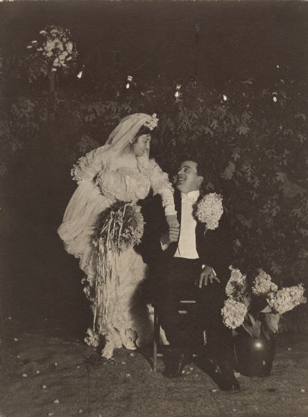 Mr. and Mrs. Louis Hobbins smile at one another and hold hands as a full-length wedding portrait is taken. She is standing, and he is sitting in a chair on a carpet spread on the ground. The bride is holding her train and bouquet in her right hand. The groom is wearing an enormous hydrangea boutonniere, perhaps as a gag. A vase of hydrangeas are in a vase on the ground next to the groom's chair. Another bouquet upper left background near a string of lights.