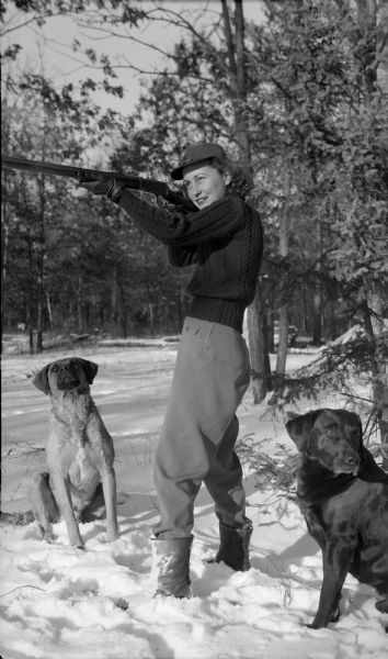 Former First Lady Dorothy Knowles hunting with two dogs. She is posed aiming her gun. There is snow on the ground and trees in the background.