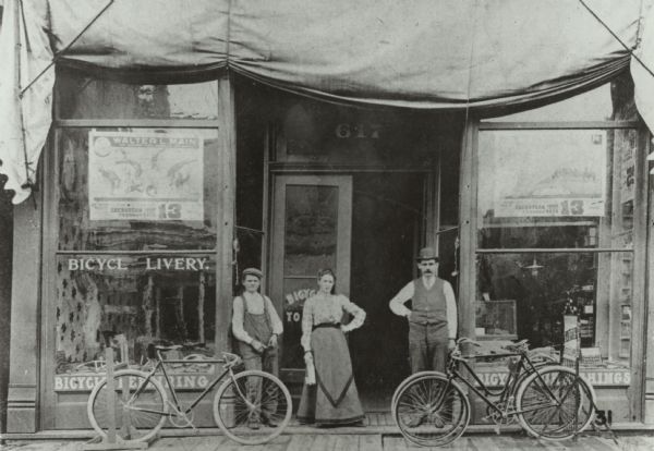 Two men and a woman pose on a wooden sidewalk in front of a bicycle livery with three bicycles. An awning is folded above the storefront, and signs and merchandise are displayed in the show windows. From left to right the people are Sammy Hanson, a young employee, Mae Tifft, and Stephen A. Tifft, owner of the livery and husband of Mae. The livery building was in an alley off of North Eighth Street and was eventually replaced by a Citizens State bank.
