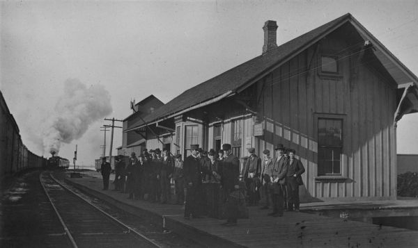 View across railroad tracks towards passengers, a large group of men and one young girl, waiting on the platform at the train station for the train to arrive. Many of the men are holding luggage. A man in a uniform at the front stands next to trunks. One train is stopped on the tracks on the left and another train is arriving on the tracks next to the platform. Signs on the station read, "Colfax," Agency National Express Co.," "Western Union Telegraph Office" and "Cap Sheaf Soda." A grain elevator and more railroad cars are behind the platform.
