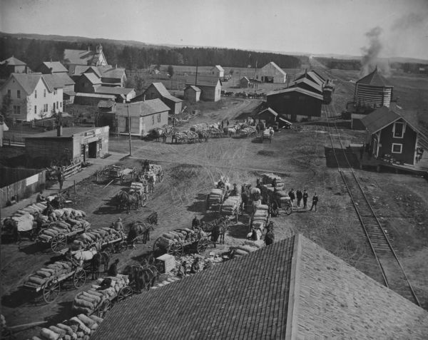 Elevated view of the railroad depot taken from the wooden shake roof of a building. On the right are railroad tracks, depot buildings, grain storage and a train sitting on the tracks. Several groups of people wait for the train to pickup their horse-drawn wagon loads of potatoes. On the left commercial buildings, residential homes and a church. A sign for Bull Durham tobacco hangs above the door to what appears to be a wagon repair business. In the background are wooded hills.