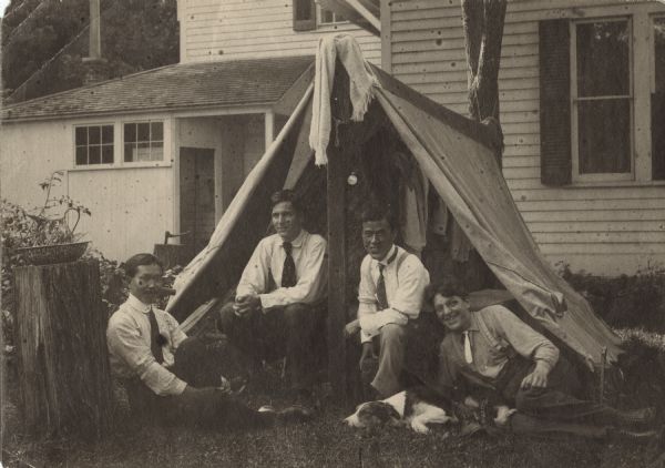 Arnold Gesell (2nd from left) and three friends posing in the open doorway of a tent, with a dog resting in the grass. The men are wearing trousers, shirts and neckties. The tent is anchored to a tree in the background, and a house is behind the tent. A towel is hanging on the peak of the tent, with a pocket watch below, with more clothing hanging inside. A tree stump on the left has an enamelware pitcher and bowl set placed on it.