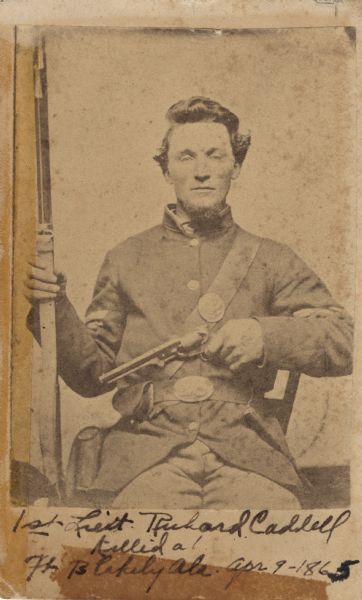 Carte-de-visite studio portrait of 1st Lieutenant Richard Cadell seated in a chair and wearing his military uniform. He holds a rifle in his right hand and a pistol across his chest in his left hand. He served in Company D, 11th Wisconsin Infantry Regiment, and was killed in the Battle of Blakely, at Fort Blakeley, Alabama, on April 9th, 1865. At the time of his death, he was a 2nd Lieutenant. It was the last major ground battle of the Civil War, and took place after the surrender of General Robert E. Lee in Virginia.